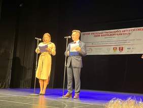Pokaż zdjęcie: Kobieta i mężczyzna stoją na scenie Białostockiego Teatru Lalek z mikrofonami i kartkami w dłoniach podczas otwarcia XXVIII Przeglądu Twórczości Artystycznej Osób Niepełnosprawnych. W tle widoczny baner z nazwą wydarzenia i logotypami organizatorów.