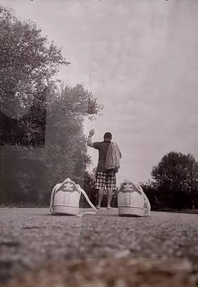 Pokaż zdjęcie: zdjęcie czarno-białe przedstawia buty a za nimi w tle stojącego tyłem na boso mężczyznę z podniesioną ręką