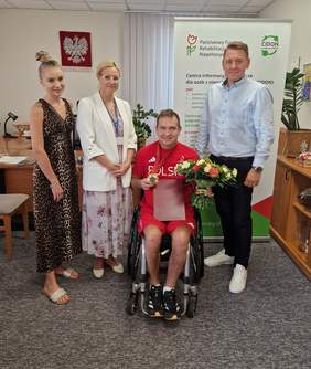 Pokaż zdjęcie: Rafał Czuper siedzi na wózku inwalidzkim z medalem na szyi i kwiatami, otoczony przez dwie kobiety i jednego mężczyznę, którzy stoją obok niego. Zdjęcie wykonano w biurze, z tłem baneru informacyjnego PFRON.