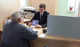 Pokaż zdjęcie: Na zdjęciu pracownik Oddziału trzyma w ręku podręcznik dla osób z niepełnosprawnością i udziela informacji klientowi. Osoby siedzą przy biurku.