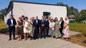 Pokaż zdjęcie: Przed białym budynkiem stoją kobiety i mężczyźni, którzy pozują do zdjęcia. Są elegancko ubrani. Dzień jest słoneczny. 