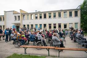 Pokaż zdjęcie: Na długich ławkach przed budynkiem stowarzyszenia siedzą goście zaproszeni na obchody jubileuszu 25 lecia. Goście mają prezenty, cukierki, kwiaty, grawertony.