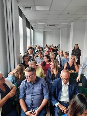 Pokaż zdjęcie: W sali na krzesłach w rzędach siedzą ludzie. Wiele osób trzyma w rękach białą laskę. 