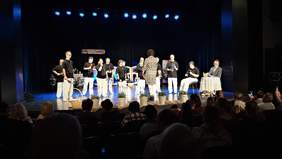 Występ na scenie artystów z Łodzi, grają na instrumentach, a z przodu (tyłem do widzów) stoi instruktorka, która dyryguje grupą