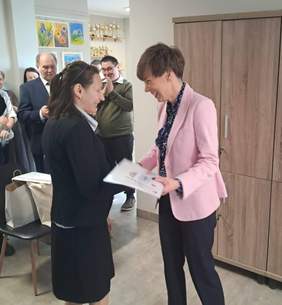 Pokaż zdjęcie: Fotografia z uroczystości przedstawiająca dwie kobiety podczas wręczania dyplomu. Jedna kobieta stoi po prawej stronie, ma krótko obcięte brązowe włosy i jest ubrana w różową marynarkę oraz ciemne spodnie; trzyma w rękach dyplom. Druga kobieta stoi po lewej stronie, ma ciemne włosy do ramion, i jest ubrana w czarną marynarkę i spódnice - jest zwrócona w stronę kobiety w różowej marynarce. W tle stoi kilka osób, które klaszczą.