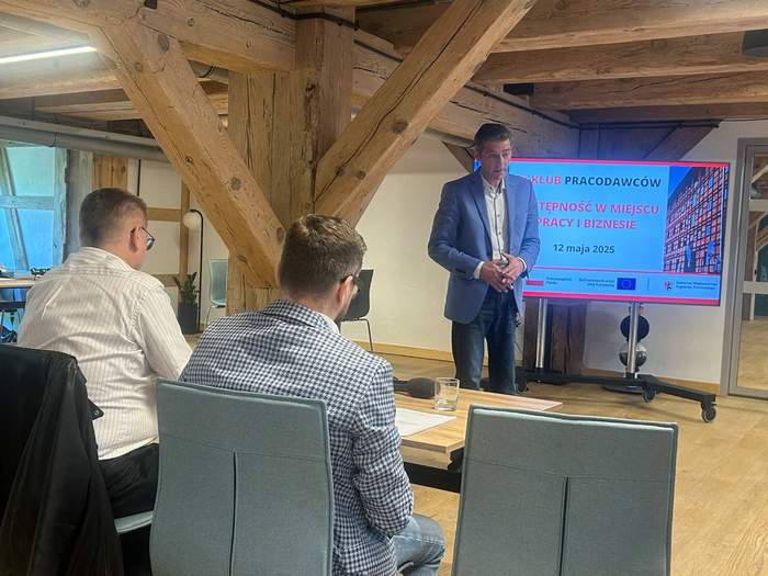 Na pierwszym planie, przy stole siedzą dwaj mężczyźni i słuchają prelegenta, który stoi przed nimi. Za mówcą widać ekran z wyświetloną prezentacją.