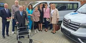 Pokaż zdjęcie: Grupa jedenastu osób stoi na parkingu, na tle dwóch busów z widoczną jedną naklejką informującą, że pojazd przystosowany jest do przewozu osób z niepełnosprawnością. Dwie osoby poruszają się o kuli, a jedna opiera się o chodzik rehabilitacyjny.