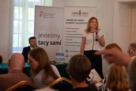 Pokaż zdjęcie: Kobieta z mikrofonem stoi w sali konferencyjnej na tle baneru Fundacji Szansa dla Niewidomych. Wystąpieniu przysłuchuje się grupa kilku osób. Za stolikiem przykrytym zielonym płótnem siedzi mężczyzna, obok którego ustawiony jest baner PFRON.