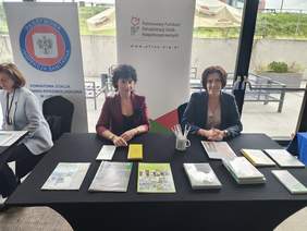 Pokaż zdjęcie: Przy stoliku na którym leżą ulotki siedzą dwie kobiety, z tyłu rollup z logo PFRON.