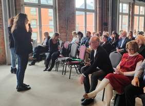 Pokaż zdjęcie: Spotkanie w nowoczesnym, loftowym wnętrzu z ceglanymi ścianami i dużymi oknami. W tle widać liczną publiczność, uważnie słuchającą kobiety przemawiającej do mikrofonu.