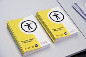 Pokaż zdjęcie: Stosy roszur "Co warto wiedzieć o dostępności". Broszury mają żółto-białą okładkę z czarnym symbolem osoby w okręgu na górze. W dolnej części okładki znajduje się tekst: "Broszura w tekście łatwym do czytania i zrozumienia", a także logotyp PFRON i innej organizacji.