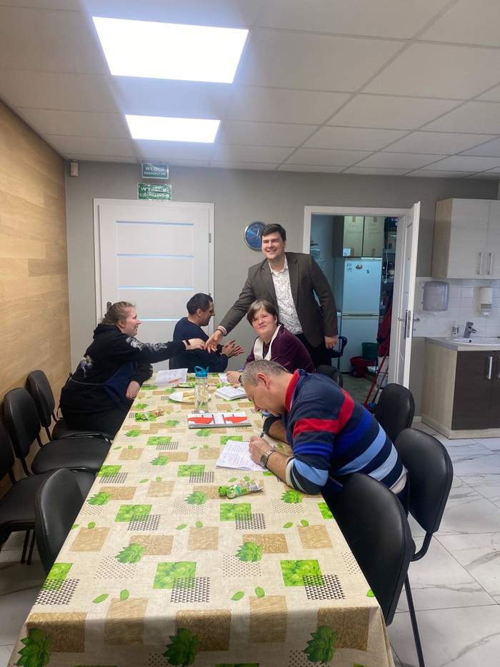 Pokaż zdjęcie: Cztery osoby siedzą przy dużym stole nakrytym beżowym obrusem w zielone liście. Jeden z mężczyzn stoi i podaje rękę siedzącej przy stole kobiecie