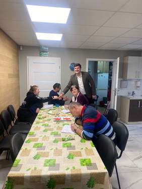 Pokaż zdjęcie: Cztery osoby siedzą przy dużym stole nakrytym beżowym obrusem w zielone liście. Jeden z mężczyzn stoi i podaje rękę siedzącej przy stole kobiecie