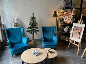 Pokaż zdjęcie: Fragment pomieszczenia w którym znajdują się dwa eleganckie niebieskie fotele, przed nimi stoi podwójny stolik z drewnianym blatem. Między fotelami stoi choinka, a z prawej strony stoi regał z różnorodnym rękodziełem i obraz na sztaludze.