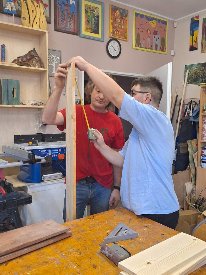 Pokaż zdjęcie: Osoba z niepełnosprawnoscią (mężczyzna) pracuje w pracowni stolarskiej z opiekunem, oboje mierzą deskę