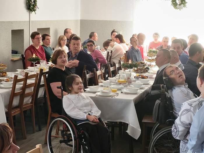 Pokaż zdjęcie: Przy wigilijmym stole siedzą osoby z niepełnosprawnościami w różnym wieku i są ubrani odświętnie oraz uśmiechnięci