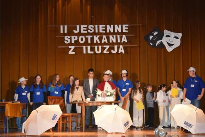 Pokaż zdjęcie: Na scenie występy grupy artystycznej udającej wywiad z papieżem Janem Pawłem II. Przed dziećmi leżą parasolki z logo TVP3.