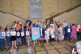 Pokaż zdjęcie: Duża grupa osób zgromadzona na schodach, przy sztaludze z plakatem konkursowym. Część z nich trzyma w dłoniach dyplomy. Przed nimi mężczyzna wykonujący fotografię. W tle banner Muzeum Narodowego z napisem Chagale.