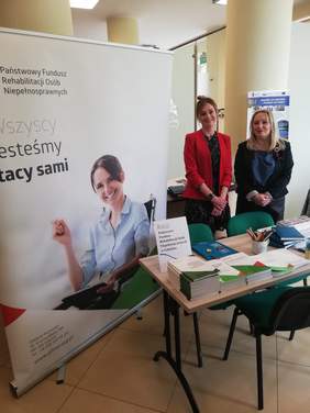 Pokaż zdjęcie: Dwie elegancko ubrane kobiety uśmiechnięte stoja przy biurku na którym leżą materiały promocyjne. Obok stoi baner z logo PFRON i zdjęciem kobiety na wóżku nwalidzkim.