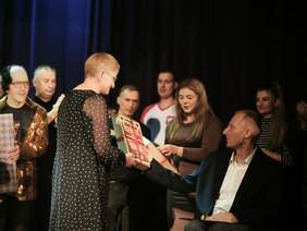 Pokaż zdjęcie: Na scenie kobieta w sukience w kropki wręcza nagrodę w czerwonym opakowaniu. W tle widać osoby obserwujące ceremonię, część z nich trzyma prezenty lub oklaskuje.