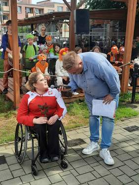 Pokaż zdjęcie: Pani siedzi na wózku inwalidzkim. Obok niej stoi pan, który zadaje jej pytania. Trzyma w rękach mikrofon i pochyla się nad panią. 