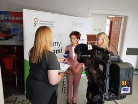 Pokaż zdjęcie: Prezes Marlena Malag stoi na tle ścianki z logiem PFRON. Z lewej strony stoi dziennikarka z mikrofonem w reku i zadaje pytania. Z prawej kamera telewizyjna. 