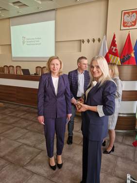 Pokaż zdjęcie: Na pierwszym planie dwie kobiety ubrane w ciemne garnitury w tle mężczyzna.