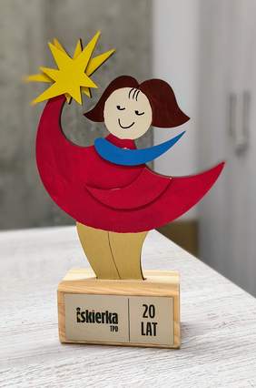 Pokaż zdjęcie: Na zdjęciu znajduje się kolorowa, drewniana statuetka przedstawiająca stylizowaną postać trzymającą żółtą gwiazdę. Postać ma na sobie czerwoną sukienkę, a wokół szyi widoczny jest niebieski szalik. Podstawa statuetki jest wykonana z jasnego drewna, a na niej znajduje się tabliczka z napisem "Iskierka TPD 20 LAT." Tło zdjęcia jest rozmyte, z widocznymi szafkami po prawej stronie i jasnym, teksturowanym blatem, na którym stoi statuetka.
