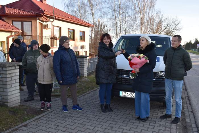 Pokaż zdjęcie: Na dworze przed siedzibą WTZ zebrała się grupa osób, jedna kobieta przekazuje drugiej kluczyki do auta. Za plecami kobiet stoi biały bus.