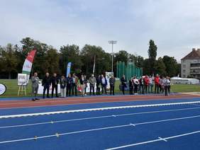 Pokaż zdjęcie: Na stadionie lekkoatletycznym na tle murawy, a wcześniej bieżni w kolorze niebieskim stoją w rzędzie obok siebie osoby biorące udział w uroczystym otwarciu zawodów