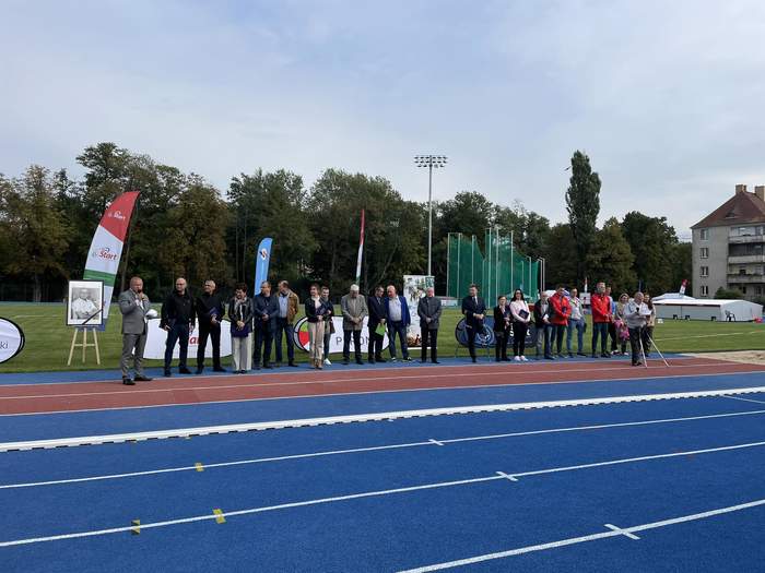 Pokaż zdjęcie: Na stadionie lekkoatletycznym na tle murawy, a wcześniej bieżni w kolorze niebieskim stoją w rzędzie obok siebie osoby biorące udział w uroczystym otwarciu zawodów
