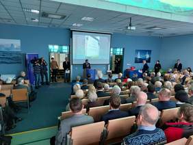 Pokaż zdjęcie: Zdjęcie z tyłu sali pokazowej w Muzeum Badań Polarnych, przy mównicy stoi mężczyzna w garniturze, po jego lewej stronie stoi dwóch kamerzystów.  