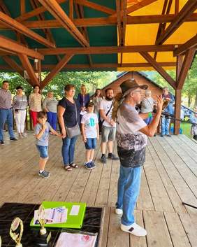 Pokaż zdjęcie: Piknik integracyjny z okazji Międzynarodowego Dnia Głuchych. Drewniana scena. Na scenie stoją dorośli i dzieci. Prowadzący wręcza upominki.