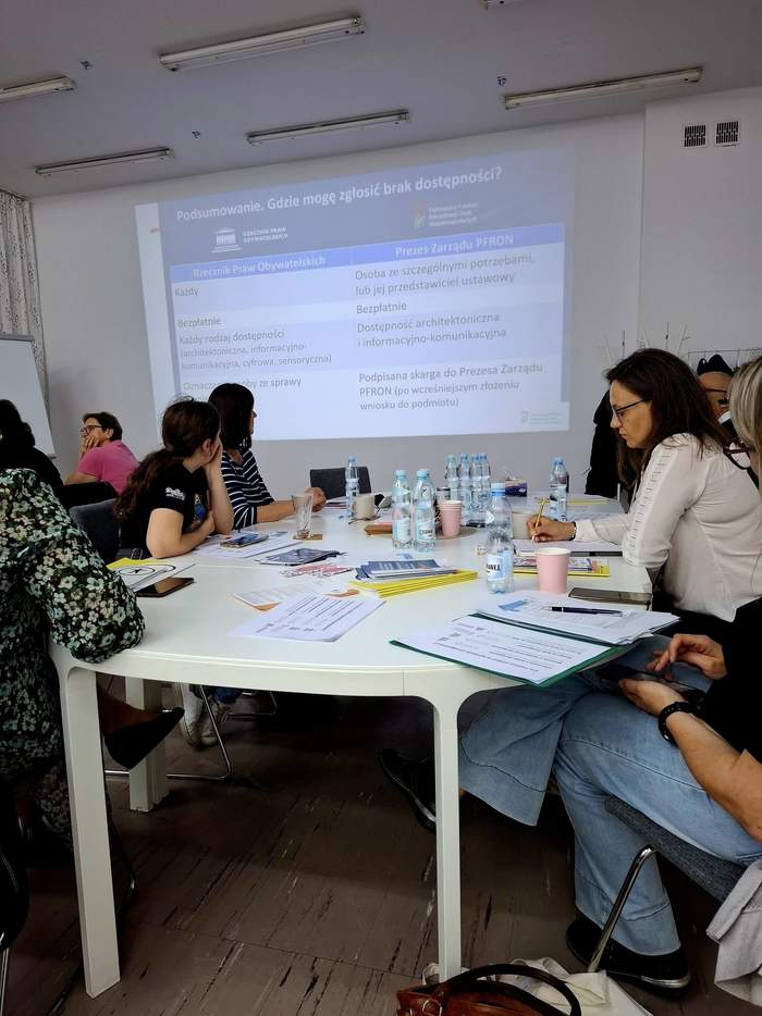 Grupa osób siedząca przy stoliku, w tle slajd z informacjami o dostępności