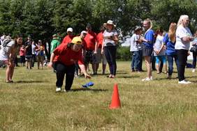 Pokaż zdjęcie: Dziewczynka biegnie po trawie podnosi woreczek i przenosi w inne miejsce, za nią stoi grupa osób, które czekają na swoją kolej w konkurencji sportowej