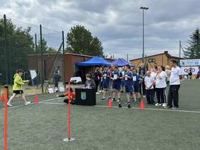 Pokaż zdjęcie: Dwie drużyny jedna w granatowych strojach sportowych druga w białych koszulkach stoją na boisku przed biała linią. Obok biurko za którym siedzi sędzia. Przez tor z boku idzie zawodniczka.