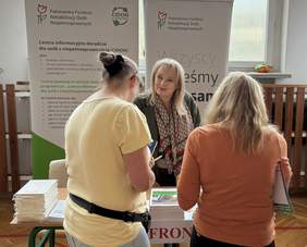 Pokaż zdjęcie: Kobieta przy stoisku Państwowego Funduszu Rehabilitacji Osób Niepełnosprawnych rozmawia z dwiema uczestniczkami, w tle widoczne banery informacyjne programu CIDON dotyczącego wsparcia dla osób z niepełnosprawnościami.