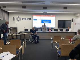 Pokaż zdjęcie: Na środku sali konferencyjnej siedzi mężczyzna na wózku inwalidzkim treningowym. Za nim długi stół prowadzących szklenie. Obok mężczyzny siedzą na krzesłach ludzie. 