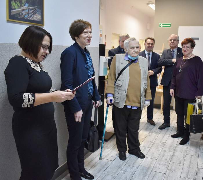Pokaż zdjęcie: Grupa pięciu mężczyzn oraz trzech kobiet stoi w korytarzu prowadzącym do poszczególnych pomieszczeń warsztatu terapii zajęciowej. Jedna z kobiet przemawia do pozostałych zgromadzonych osób posługując się materiałami, które trzyma w dłoniach. Jeden mężczyzn znajdujący się na korytarzu jest osobą, która porusza się o kuli.