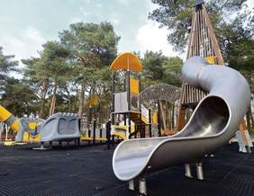 Pokaż zdjęcie: Wieża ze zjeżdżalniami, karuzela oraz inne urządzenia zabawowe na tle wysokich sosen.