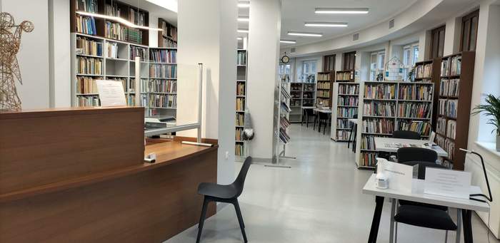Pokaż zdjęcie: Wnętrze biblioteki, po prawej regały z książkami i  biurko z krzesłem pod ścianą, po lewej lada z niskim blatem. Po środku po łuku szerokie przejście przystosowane dla osób poruszających się na wózkach inwalidzkich.