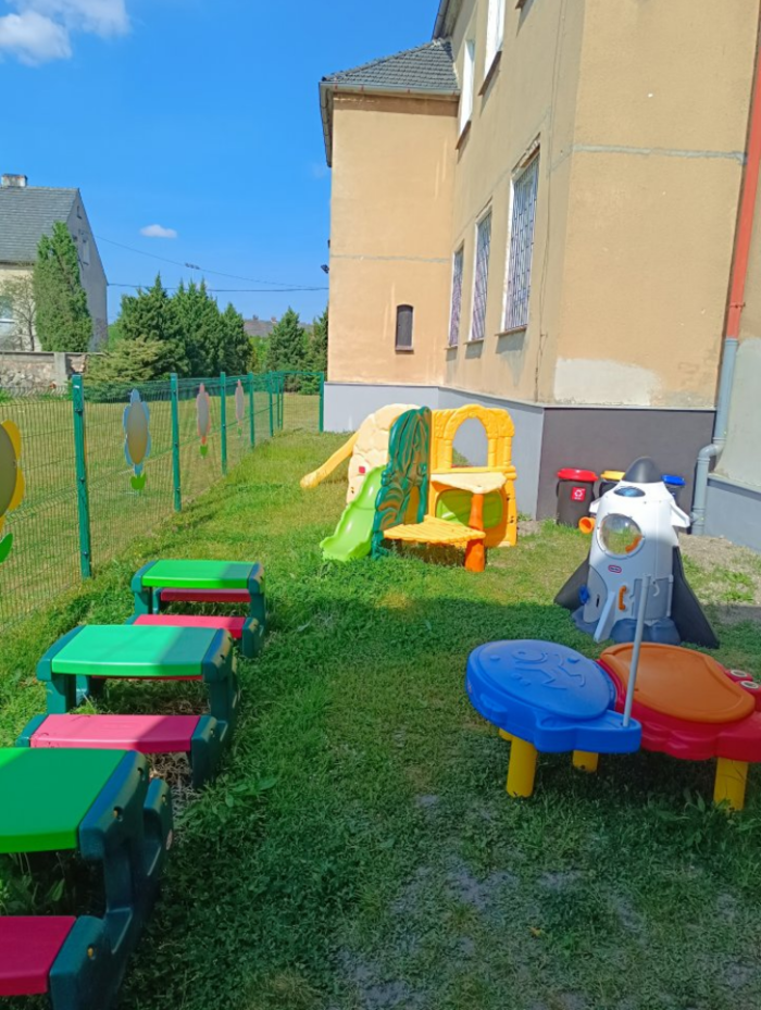 Pokaż zdjęcie: Widok na plac zabaw, widać kolorowe urządzenia, budynek przedszkola