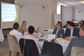 Pokaż zdjęcie: Na zdjęciu jest siedem osób w sali konferencyjnej. Za nimi wyświetlona prezentacja