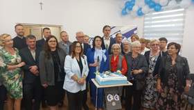Grupa osób stoi w pomieszczeniu wokół stołu z tortem ozdobionym na biało i niebiesko; w tle dekoracje z balonów, krzyż na ścianie oraz roll‑up z logo. Uczestnicy ustawieni do wspólnego zdjęcia.
