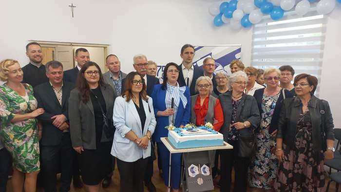 Pokaż zdjęcie: Grupa osób stoi w pomieszczeniu wokół stołu z tortem ozdobionym na biało i niebiesko; w tle dekoracje z balonów, krzyż na ścianie oraz roll‑up z logo. Uczestnicy ustawieni do wspólnego zdjęcia.