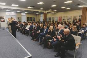 Pokaż zdjęcie: Zdjęcie przedstawia dużą, jasną salę wykładową. Po lewej stronie, na niskim podwyższeniu, stoją dwie kobiety – jedna z nich trzyma mikrofon i przemawia. Publiczność, licząca kilkadziesiąt osób, siedzi w rzędach krzeseł, zwrócona w stronę prelegentek. Uczestnicy to osoby w różnym wieku, ubrani w stroje biznesowe i casualowe; wielu z nich ma identyfikatory na szyi. W tle widać ściany z oknami oraz tablice z plakatami edukacyjnymi. Na pierwszym planie po prawej siedzi mężczyzna, który fotografuje telefonem komórkowym.