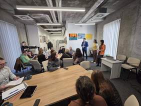 Pokaż zdjęcie: Prezentacja podczas spotkania organizacji społecznych w sali konferencyjnej. Cztery osoby stoją z przodu i przemawiają do siedzących uczestników. Na ścianach wiszą kolorowe obrazy, a uczestnicy siedzą przy stołach i słuchają wystąpienia.