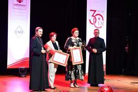 Pokaż zdjęcie: Na scenie znajdują się dwie kobiety trzymające dyplomy oraz dwaj duchowni ubrani w sutanny. W tle widoczne są banery z logo Caritas oraz informacją o 30-leciu istnienia organizacji.