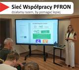Sala szkoleniowa z uczestnikami siedzącymi tyłem do aparatu przed dużym ekranem na stojaku. Nad ekranem wisi baner z logo „Sieć Współpracy PFRON” i hasłem „działamy razem, by pomagać lepiej”. Na ekranie wyświetlana jest prezentacja z napisem „OWiT Kalisz Ośrodek Wsparcia I Testów” oraz grafiką złączonych dłoni. Po prawej stronie ekranu stoi kobieta, która prowadzi spotkanie. Obok niej na małym stoliku, znajduje się otwarty laptop.