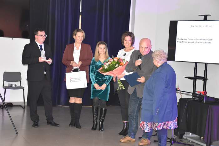 Pokaż zdjęcie: na scenie stoi 6 osób, od lewej mężczyzna w ciemnym garniturze, kobieta w brązowej marynarce, Kobieta w zielonej sukience, Kobieta w białej bluzce, która trzyma kwiaty przed sobą, mężczyzna w ciemnej marynarce, kobieta ubrana w niebieski sweter i kwiecistą spódnicę.